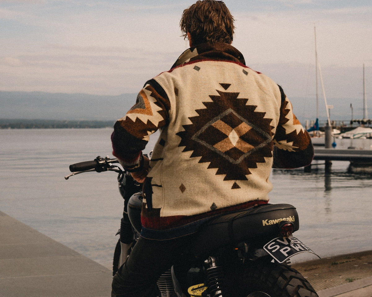 Man on a motorcycle by a lake wearing the Peguche Jacket, handwoven diamond motif across the back