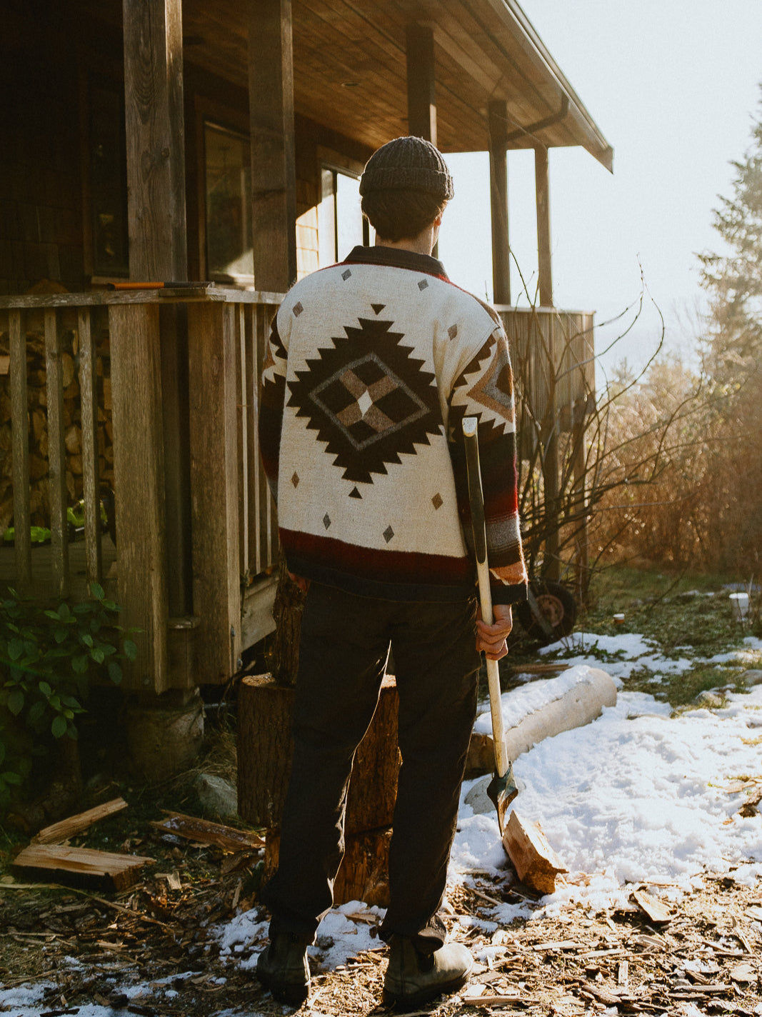 Man outside a wooden cabin in the snow wearing the Peguche Jacket, diamond motif on the back