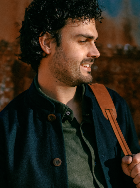 Smiling man in the Tambo Wool Jacket in Navy, holding a leather bag strap