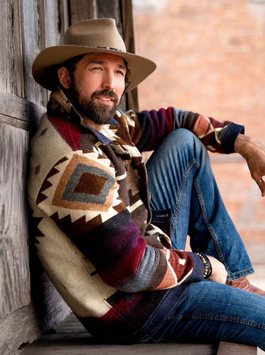 Man in a wide-brim hat wearing the Peguche Jacket, handwoven Andean wool in cream, brown and red, seated by a wooden wall